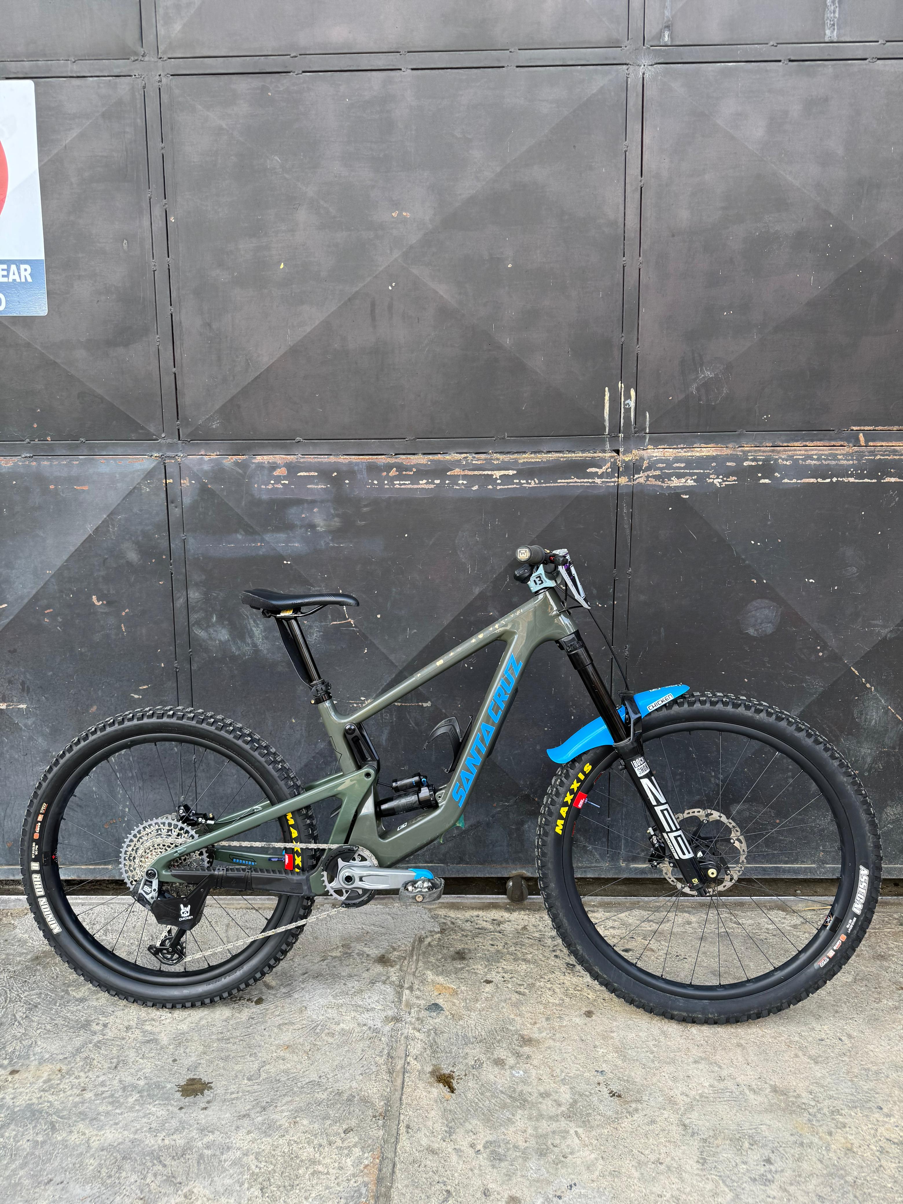 Vista lateral amplia de la bicicleta demo Santa Cruz Bronson verde oliva con detalles azules.