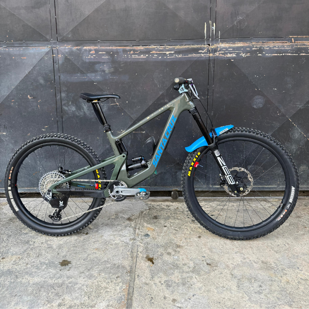 Bicicleta demo Santa Cruz Bronson verde oliva en vista lateral frente a una puerta oscura.